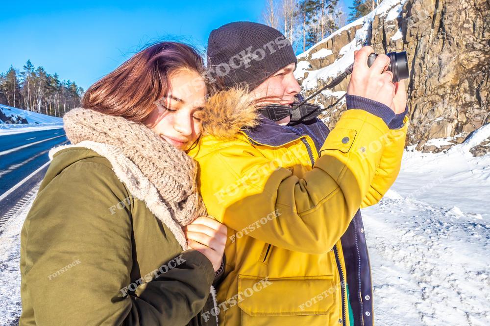На фотографии изображена женщина в теплой зеленой куртке и шарфе, прижимающаяся к мужчине в ярко-желтой куртке, который фотографирует зимний пейзаж на камеру. Фон состоит из снежной дороги, скал и голубого зимнего неба. Снимок символизирует радость совместного отдыха и красоты природы зимой.